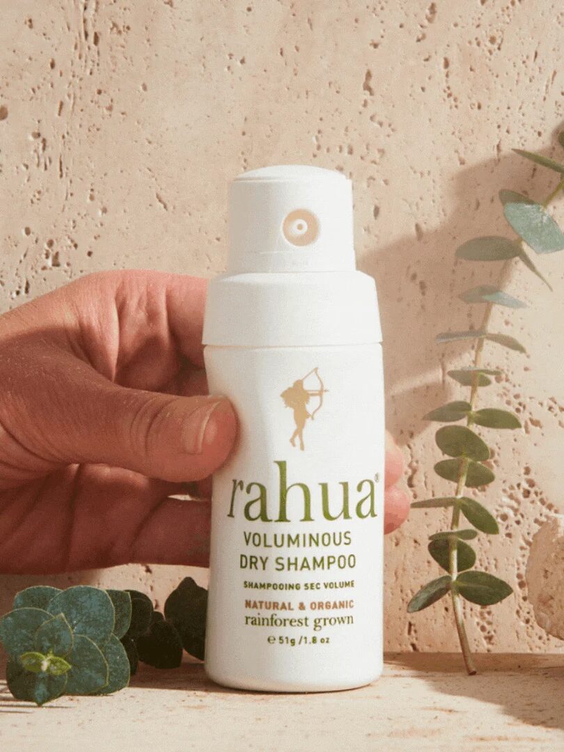 A hand holding a bottle of Rahua Voluminous Dry Shampoo against a beige textured background, with green leaves nearby.