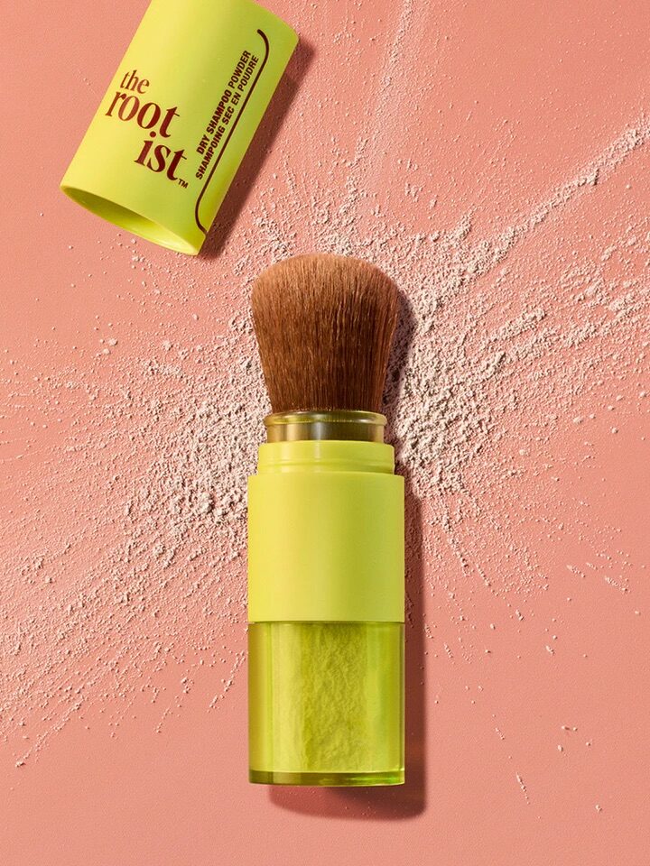 A green cosmetic brush container with an open lid lies on a pink surface with powder sprinkled around it. The container reads "the root ist.