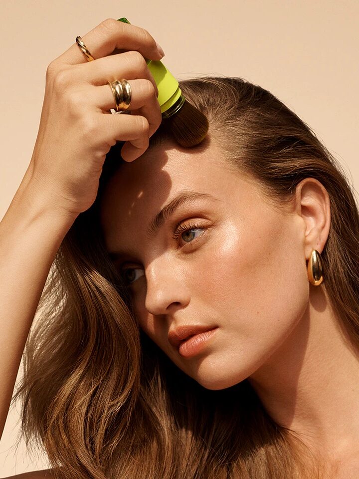 A woman with long hair applies makeup to her forehead using a brush. She is wearing several rings and gold hoop earrings.