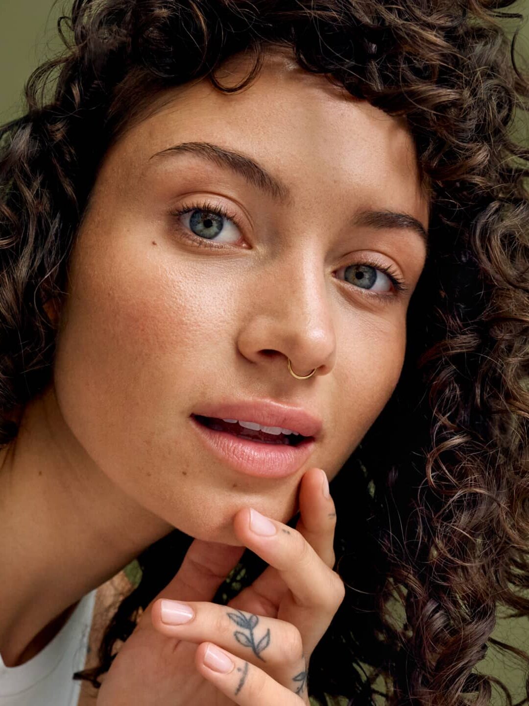 Young woman with curly hair and a nose ring gently touching her face, looking directly at the camera against a plain olive green background.