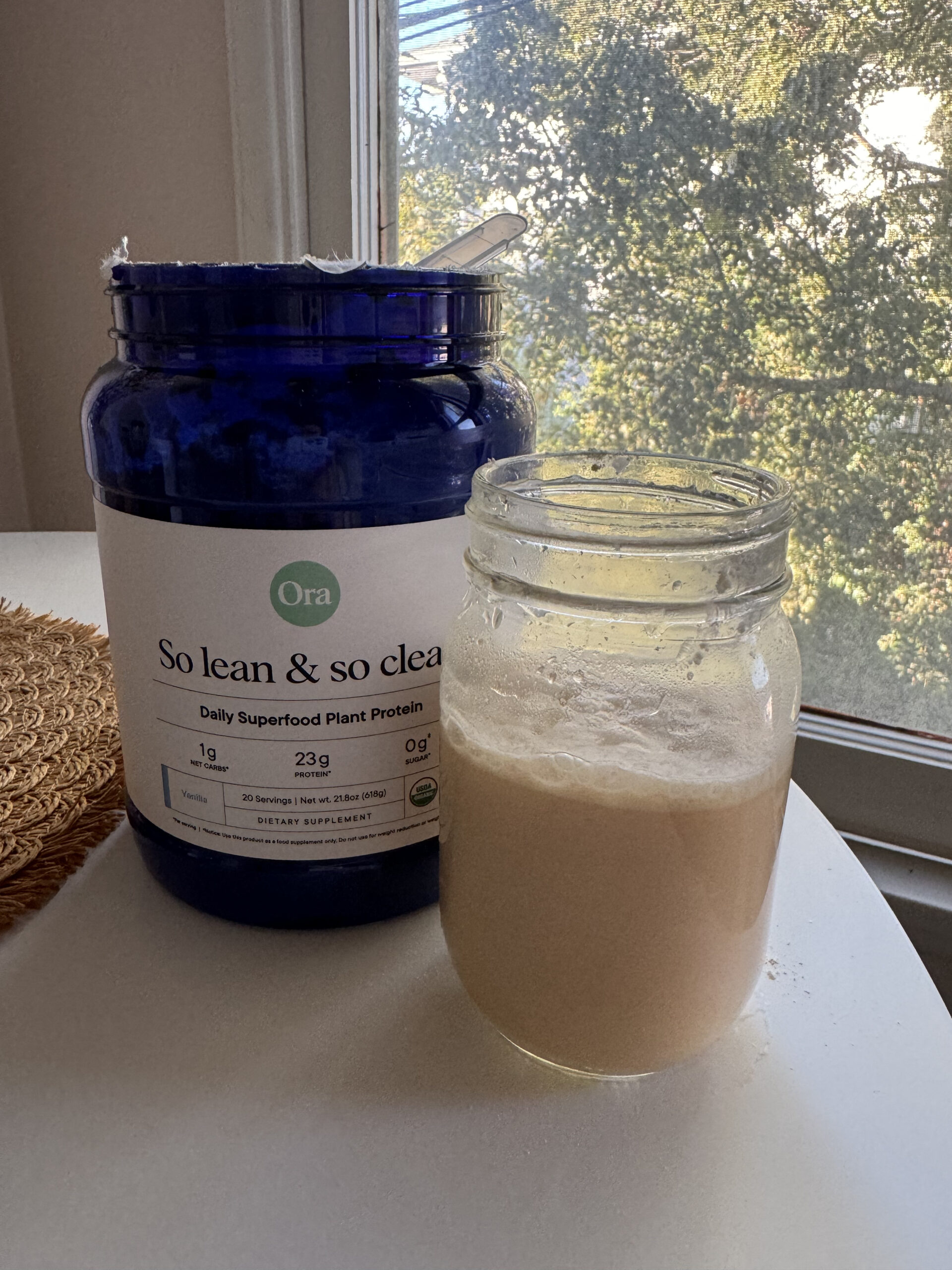 A jar of plant protein powder next to a glass of mixed drink on a table by a window.