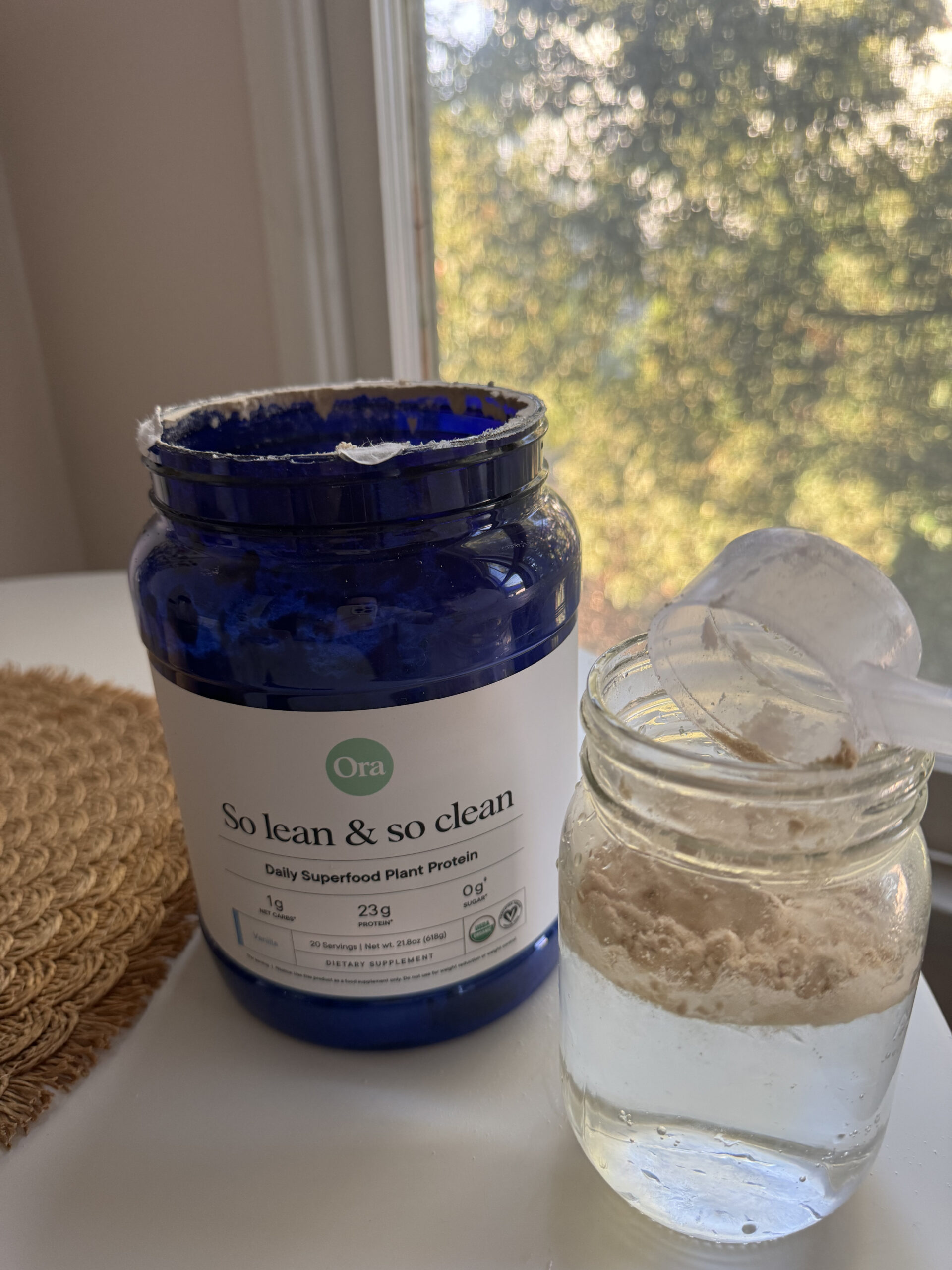 A blue container of "So lean & so clean" plant protein powder is next to a jar of water with a partially filled scoop. Background shows a window with trees.