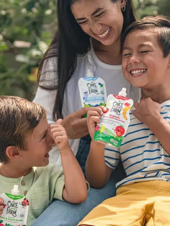 A woman sits outdoors with two boys, all smiling and holding food pouches. The boys appear to be laughing, while the woman looks at them affectionately. Trees and a fence are visible in the background.