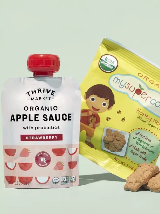 A pouch of Thrive Market organic apple sauce with probiotics and strawberry flavor is next to a package of MySuperCookies organic honey heroes whole grain cookies against a light green background.