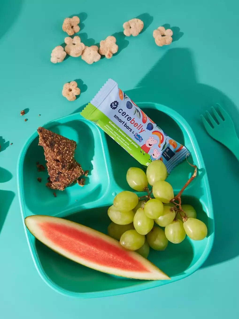 A turquoise background with a child's divided tray containing grapes, watermelon, a granola bar, an apple sauce pouch, a peeled banana, a bowl of blueberries, a granola bar, and a wrapped snack bar.