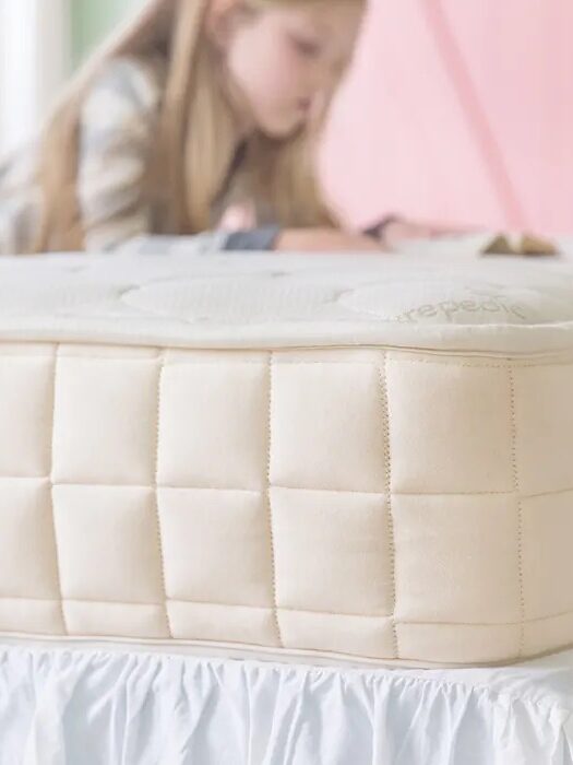 A child with long hair is laying on a bed, reading a book. The focus is on the beige, quilted mattress with a white bed skirt underneath.