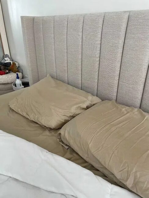 Two beige pillows rest on a matching bedspread, partially covered by a white blanket. A light-colored, upholstered headboard is behind the bed. A small collection of toys is visible on the left side.