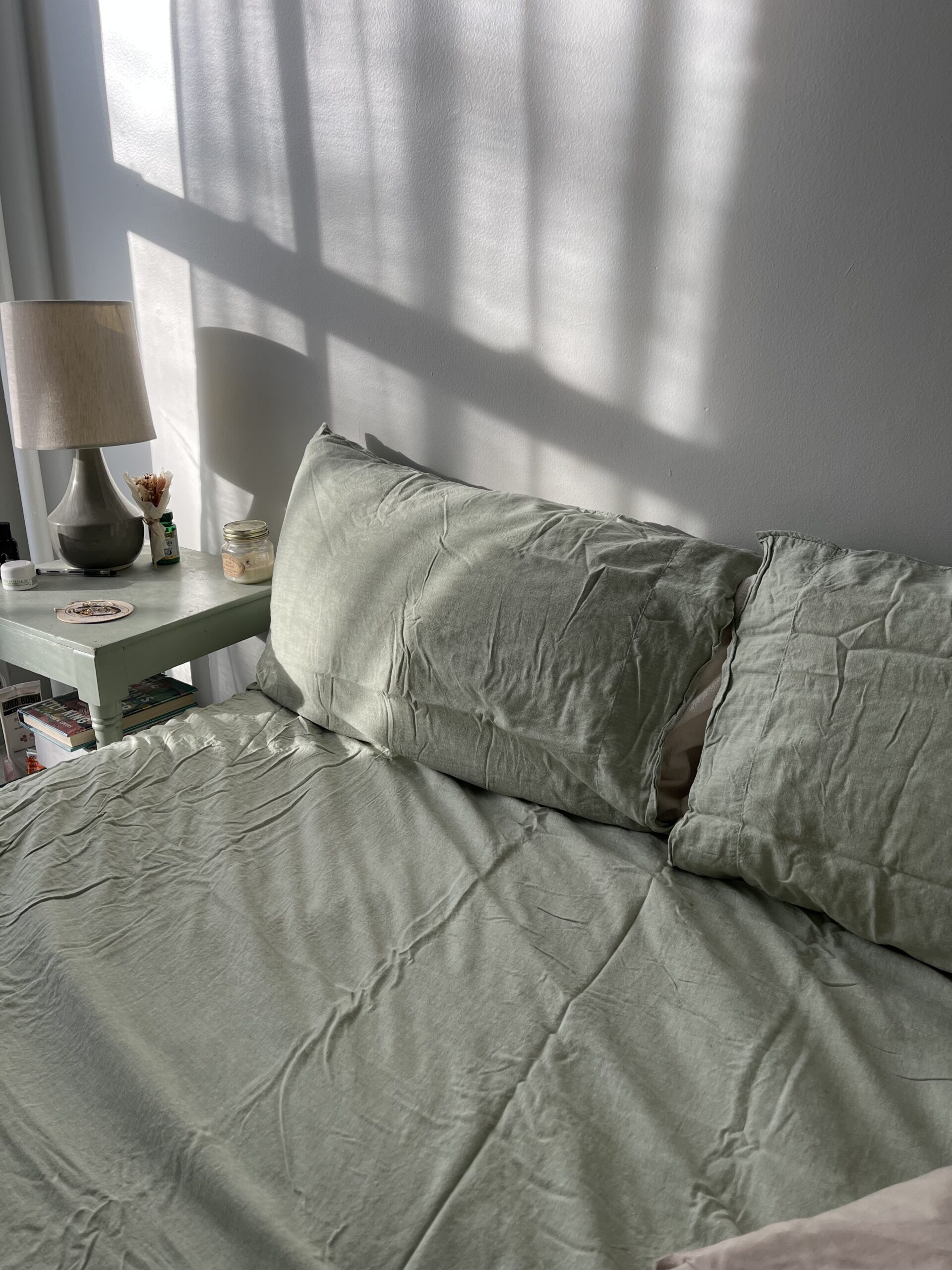 A neatly made bed with grey-green sheets next to a white bedside table, featuring a lamp, a jar, and a few small items. Sunlight casts shadows on the wall and bed.