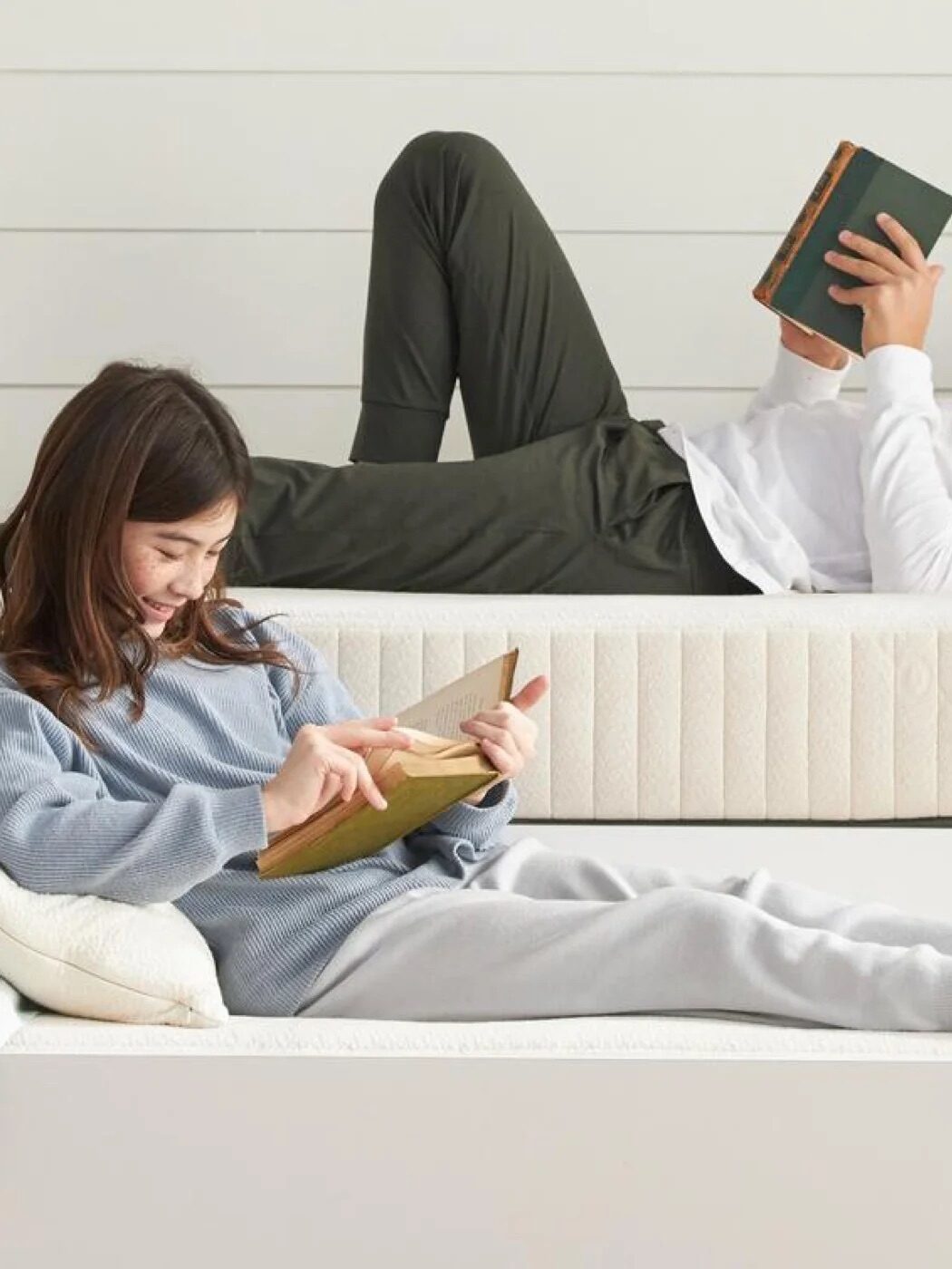 Two people are lying on mattresses, reading books. The person in the foreground is wearing a blue sweater and grey pants, while the person in the background is in a white shirt and green pants.