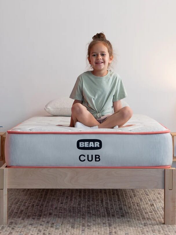 A child sits cross-legged on a bed in a minimalist room with a lamp, books, toys, and a soccer ball on the floor. The bed has a mattress labeled "Bear Cub".