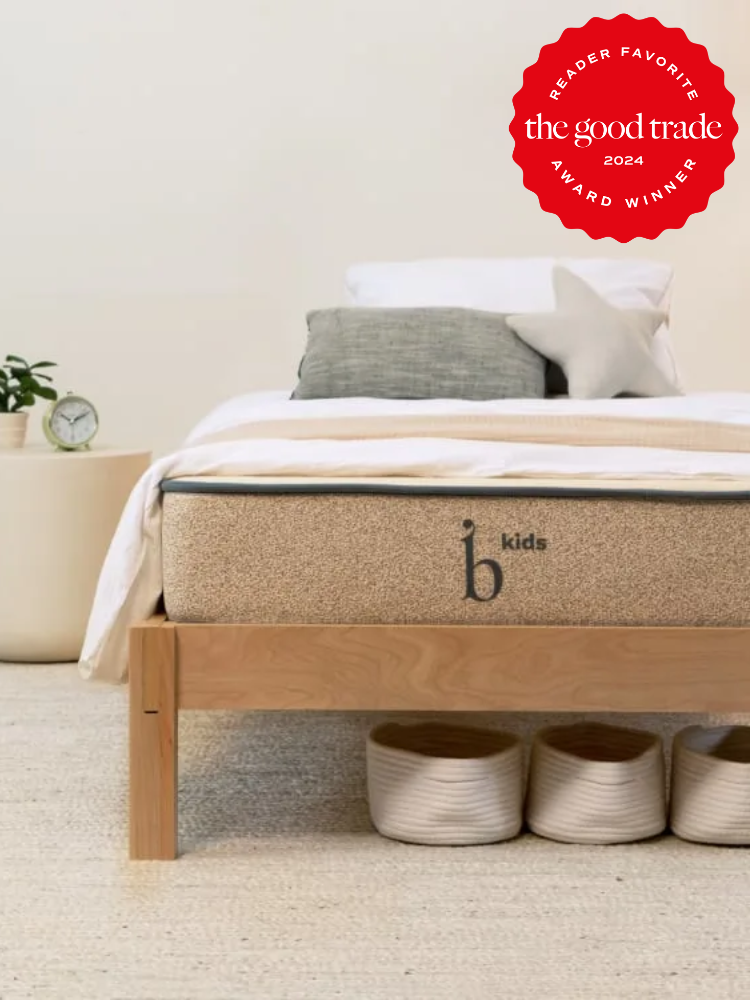 A neatly made bed with a beige mattress labeled "b kids" is displayed, with an award sticker on the upper right reading "Reader Favorite, the good trade, 2024 Award Winner." Three baskets sit under the bed.