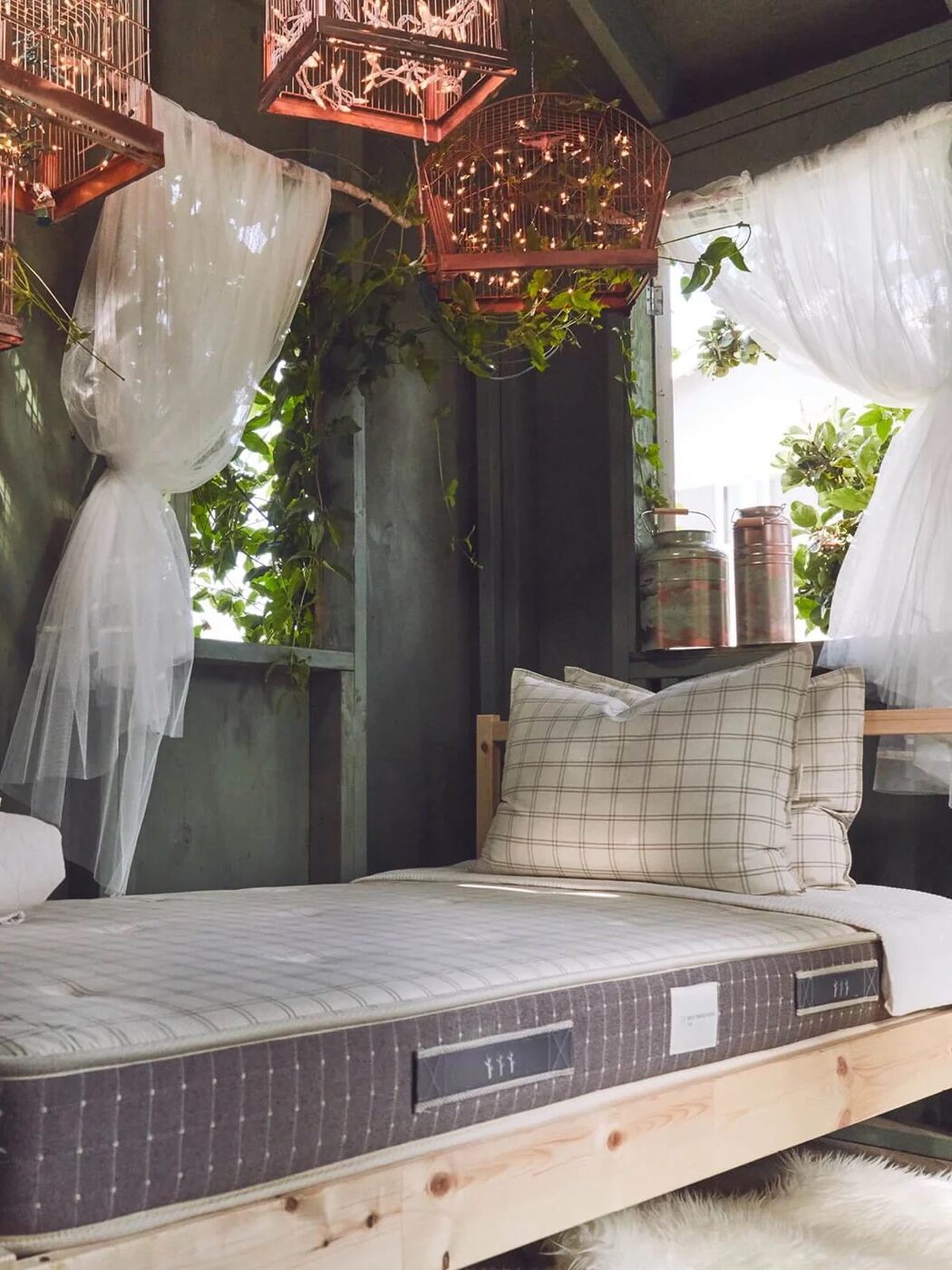 A bed with a checkered pillow and folded blankets is placed near a window with sheer curtains. Above, there are metal birdcages with hanging lights. Green plants are visible through the window.