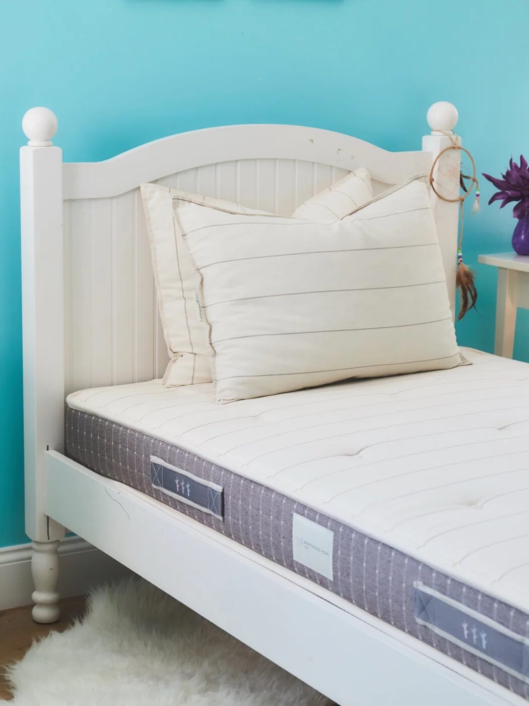 A neatly made bed with white and beige linens, a mattress, and pillows, set against a light blue wall. The adjacent white nightstand holds an alarm clock and a purple flower in a vase.