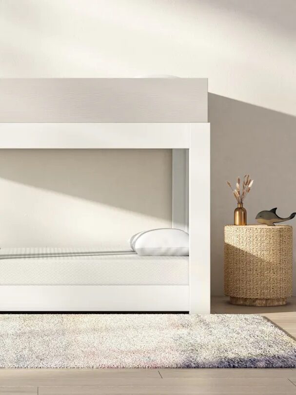 Minimalist white bunk bed with a ladder and shelves, placed in a bright room with light-colored walls, a gray rug, and a woven basket on the right. GREENGUARD Gold certification label on the upper left.