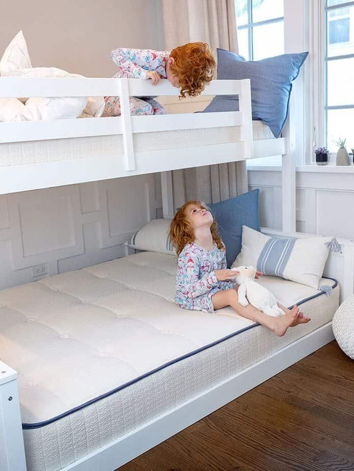 Two children, dressed in pajamas, interacting on a white bunk bed in a bright room with wooden floors and potted plants on the window sill.