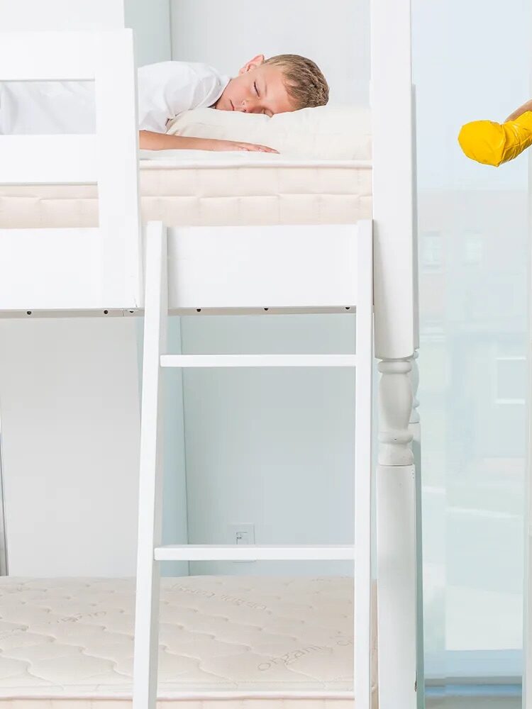 A child sleeps on the top bunk of a white bunk bed next to a bookshelf filled with colorful books and some stuffed animals.