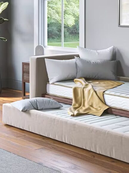 A room with a trundle bed, wooden dresser, potted plant, and a large window. The trundle bed has a beige frame and is positioned under a daybed with pillows and a blanket.