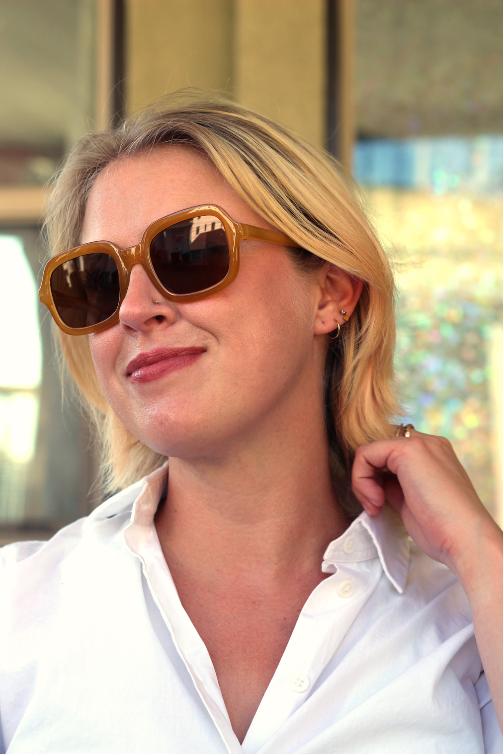 Woman wearing large brown sunglasses and a white button-up shirt, smiling slightly while touching her ear. Background is blurred.