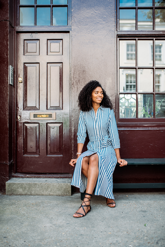 A woman in a striped dress sits on a ledge in front of a wooden door.