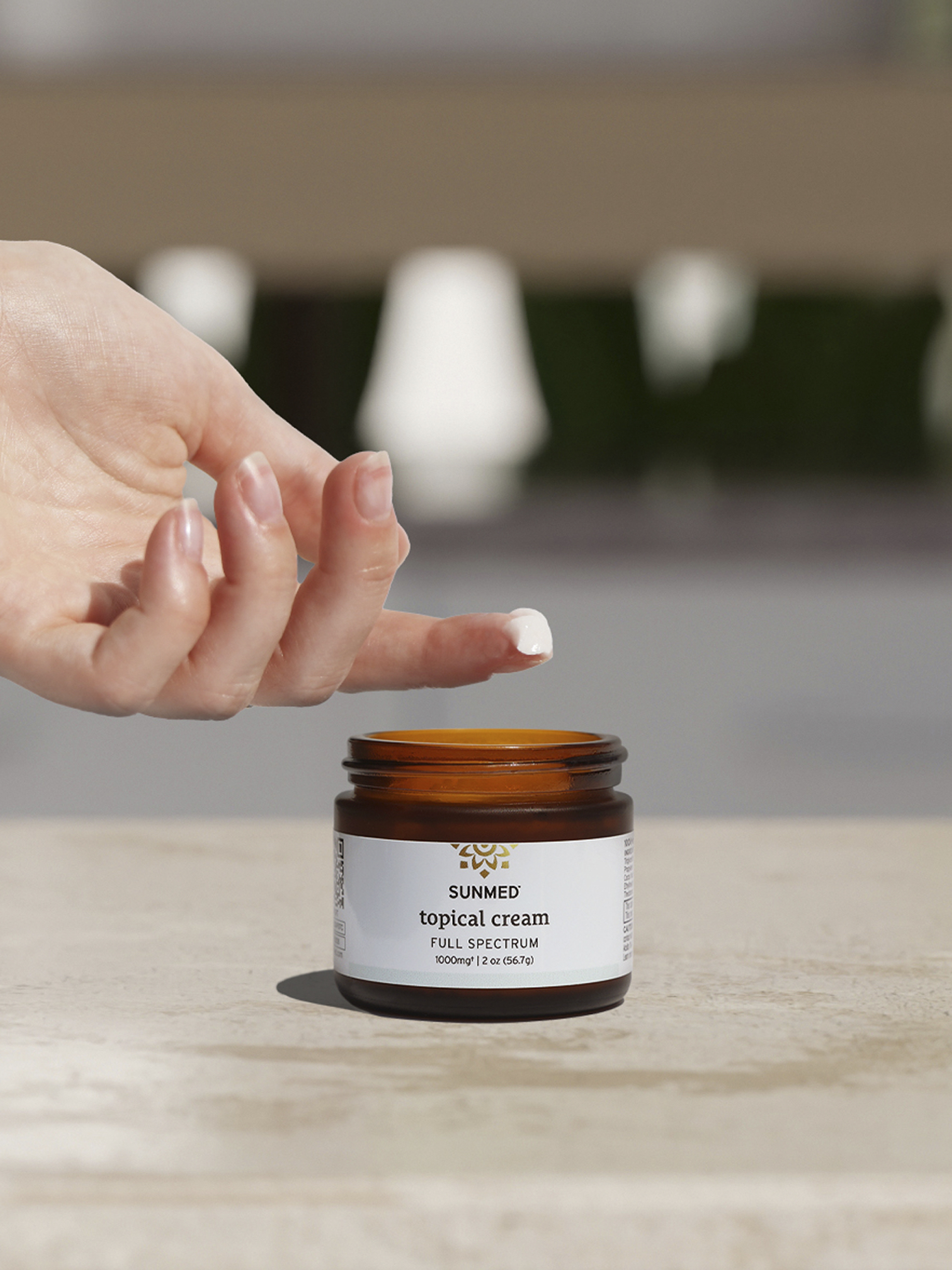 A hand is shown reaching towards an open jar of Sunmed Topical Cream Full Spectrum placed on a flat surface.