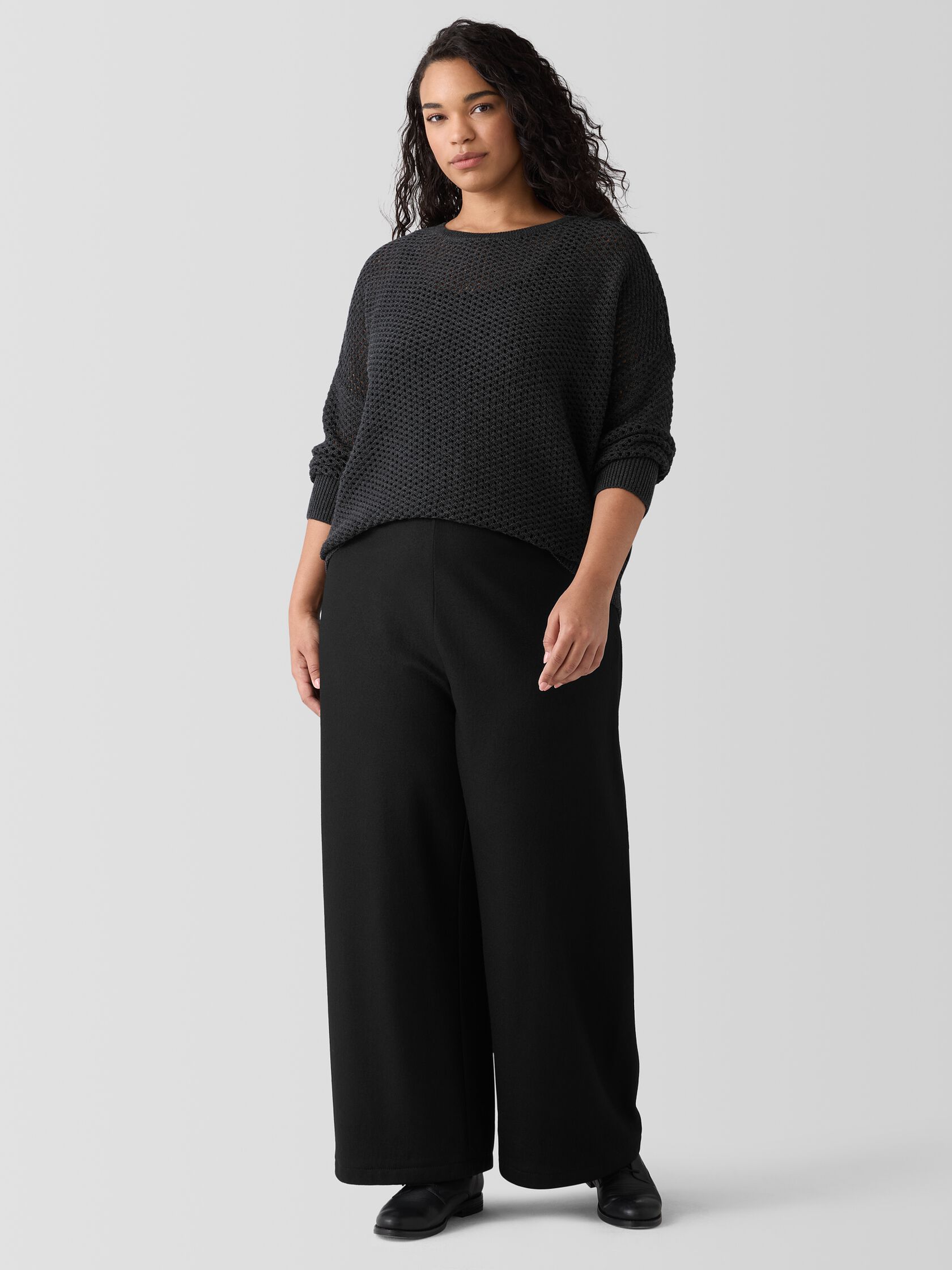 A plus size model wearing a charcoal crew neck sweater and black wide leg dress pants from Eileen Fisher. 