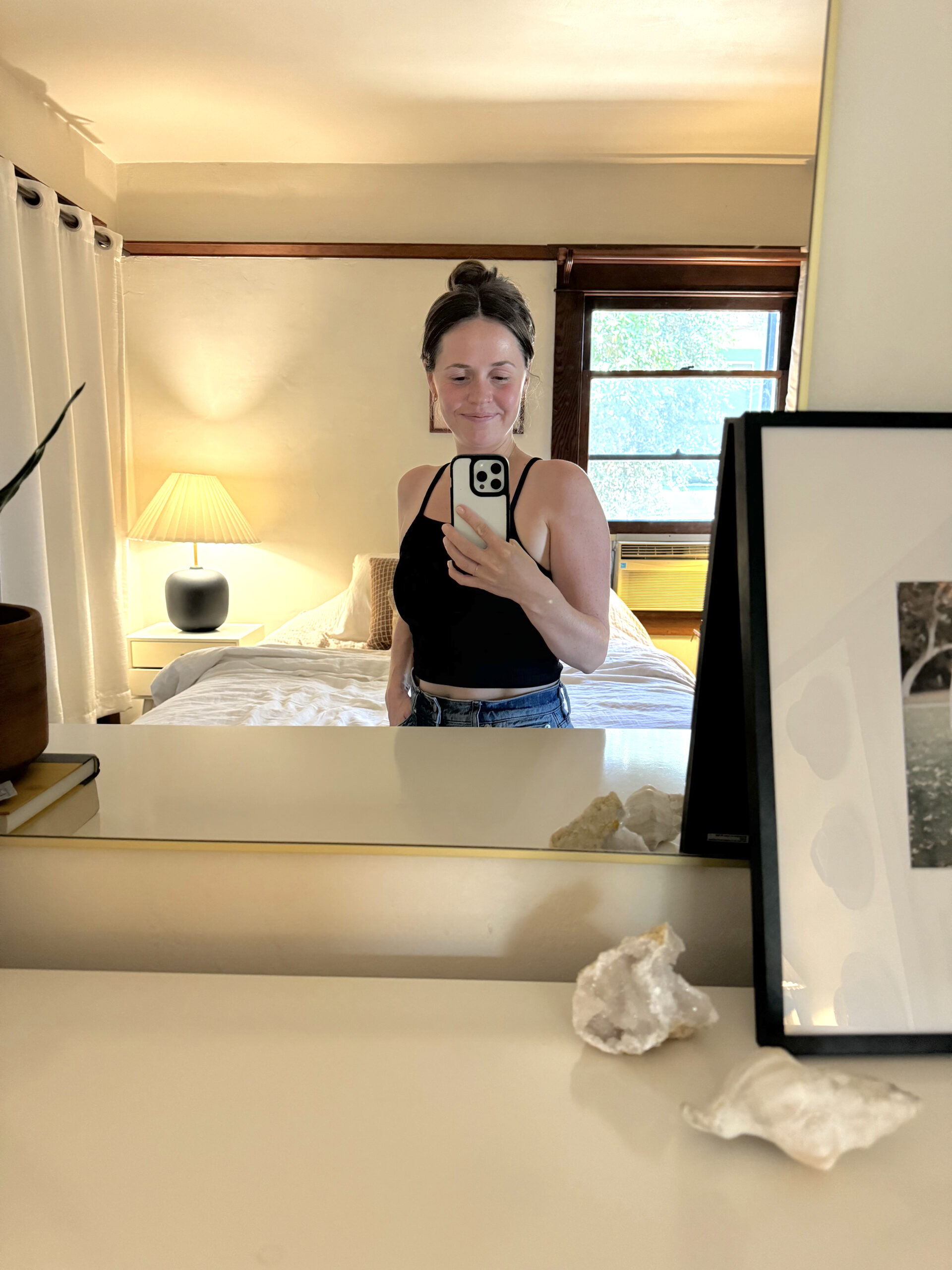 A woman takes a mirror selfie in a bedroom with a neatly made bed, lamp, and framed picture on a dresser with decorative items.