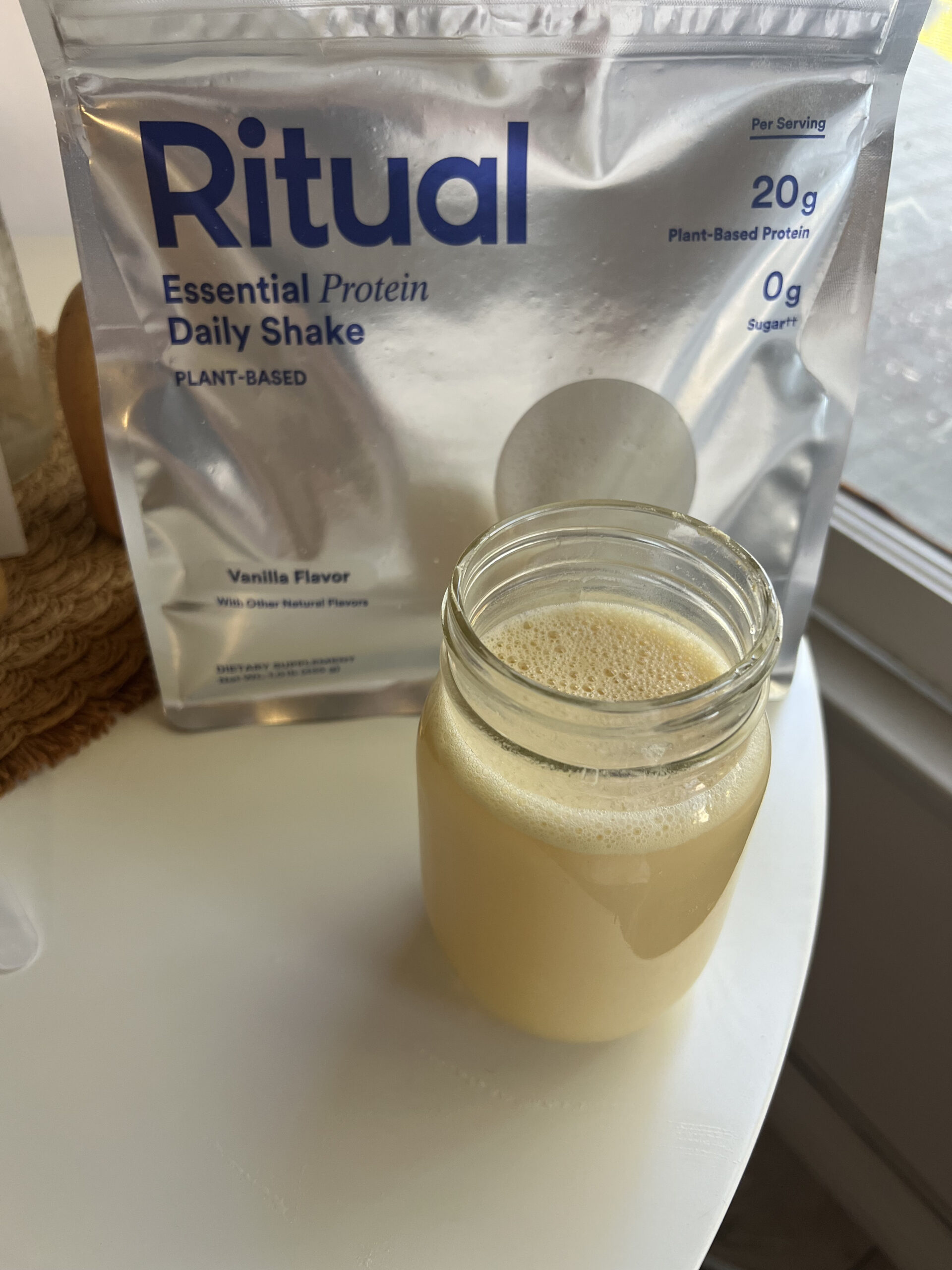 A jar of light beige protein shake is on a table in front of a bag labeled "Ritual Essential Protein Daily Shake, Vanilla Flavor.