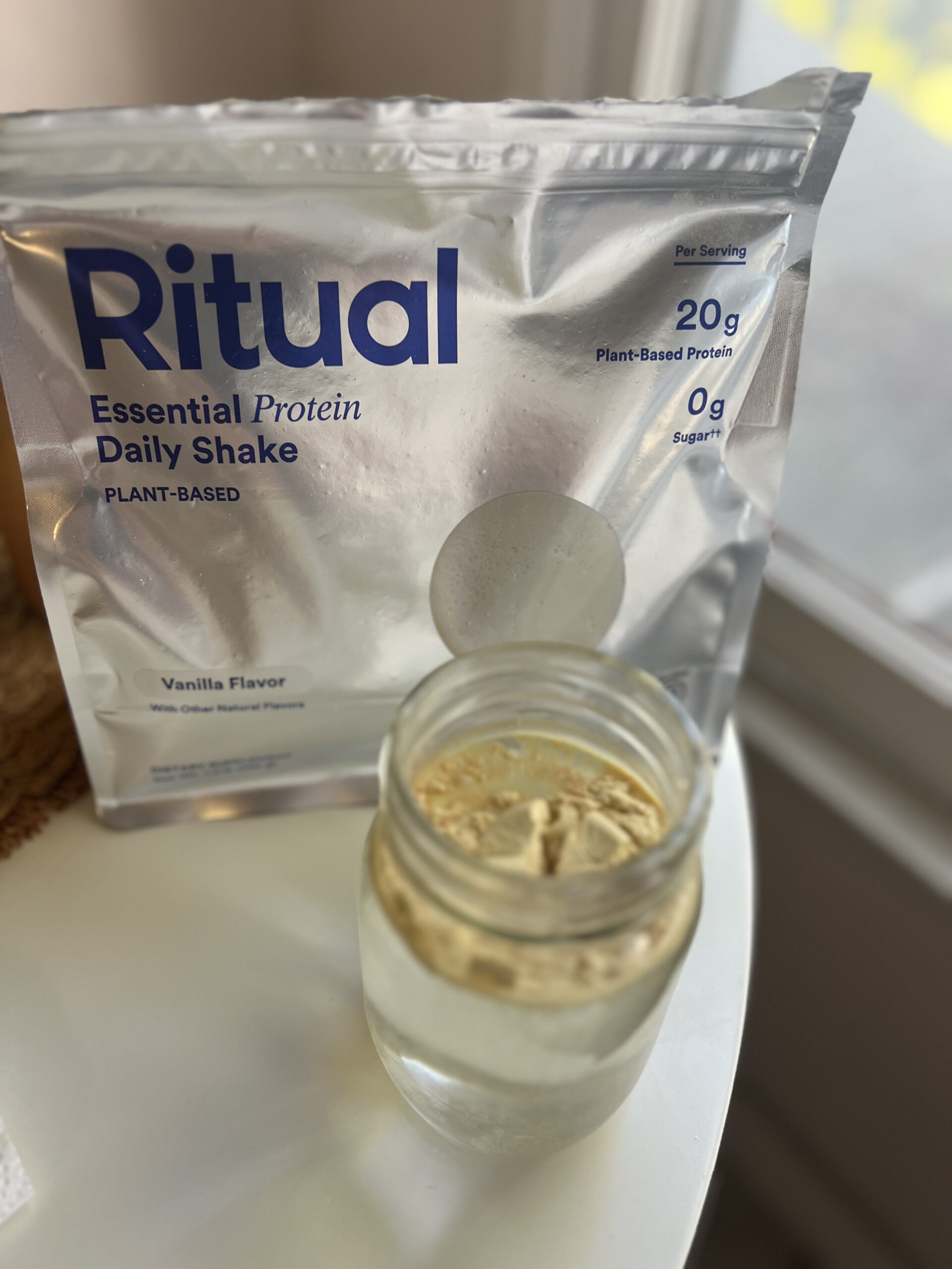 A pack of Ritual Essential Protein Daily Shake in vanilla flavor is next to a glass jar filled with powder.