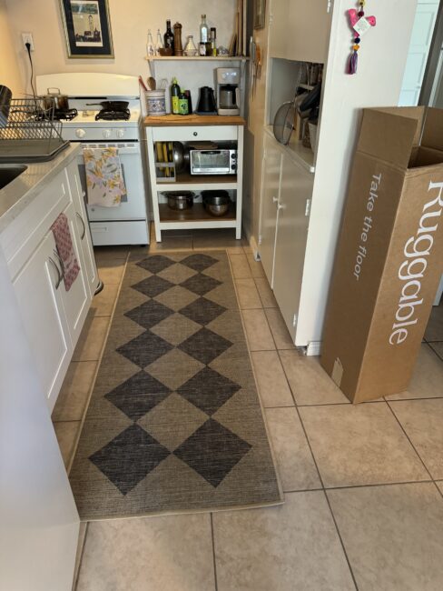 A kitchen with a stove, countertop, and shelves holding kitchen appliances. A rug with a diamond pattern is on the tiled floor, and a cardboard box labeled "Ruggable" is on the right.