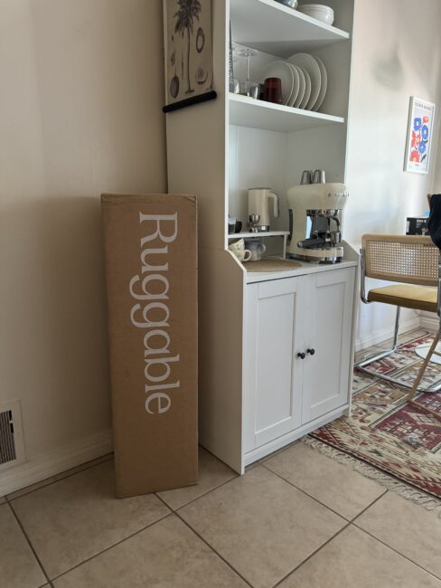 A tall Ruggable box stands next to a white cabinet in a kitchen. The cabinet holds dishes, a mixer, and a coffee maker. A patterned rug and a chair are visible on the tiled floor.