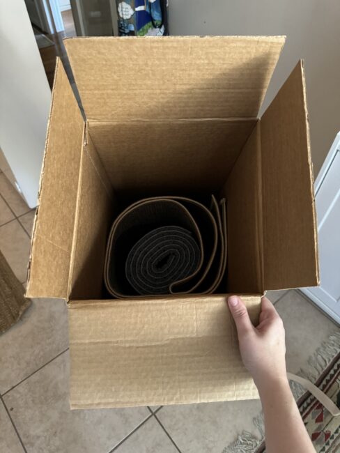 An open cardboard box containing a rolled-up mat, held by a person above a tiled floor.