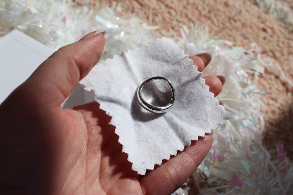 A hand holds a silver ring on a white cloth, with a textured surface in the background.