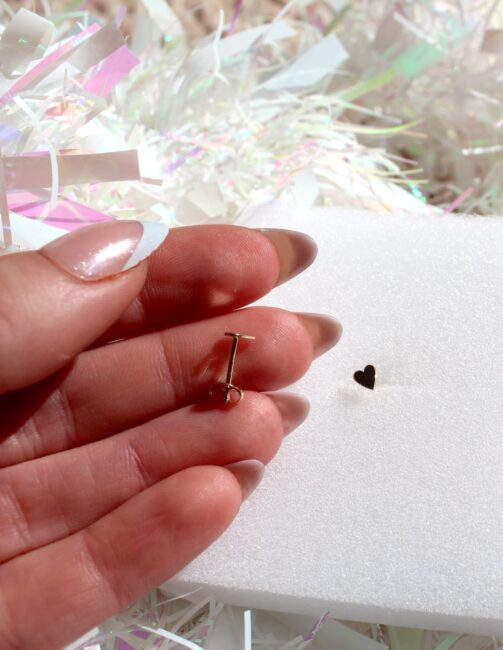 A hand holding a small gold pin above a foam surface with a heart-shaped indentation.