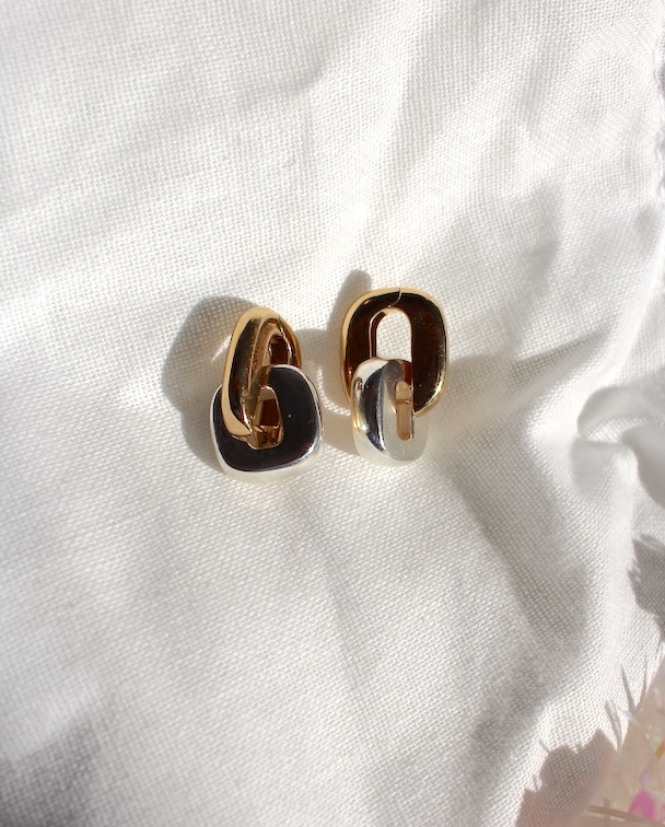 Two linked earrings in gold and silver tones resting on a crumpled white fabric surface.