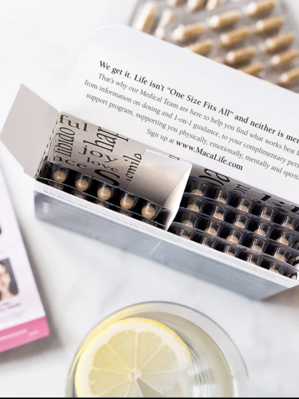 Open box of supplement capsules next to a glass of water with lemon slices and a brochure filled with text and photos.