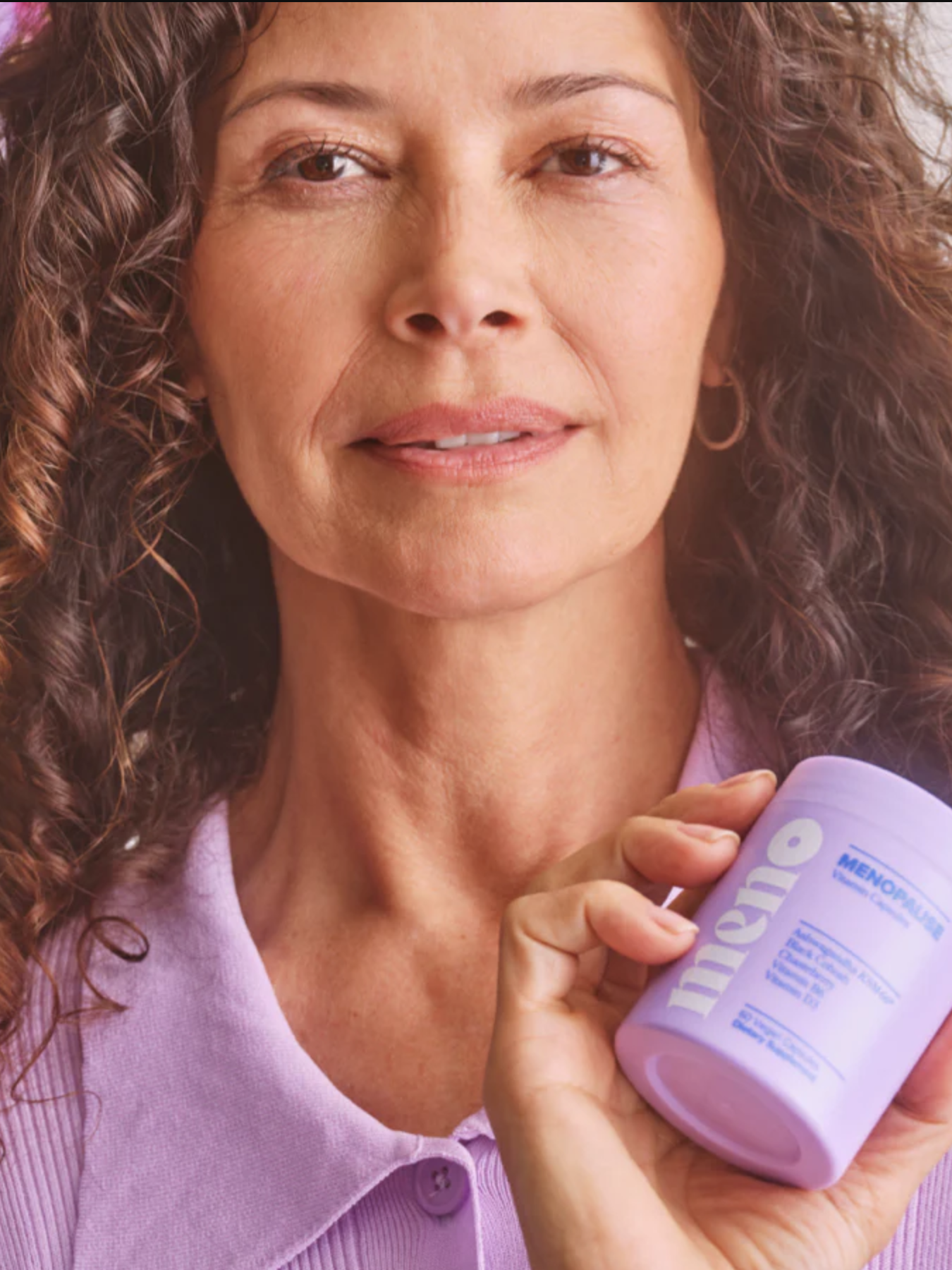 A person with curly hair wearing a lavender shirt holds a purple container of Olly's "MenoGlow" vitamins in their hand. Flowers are visible in the background.