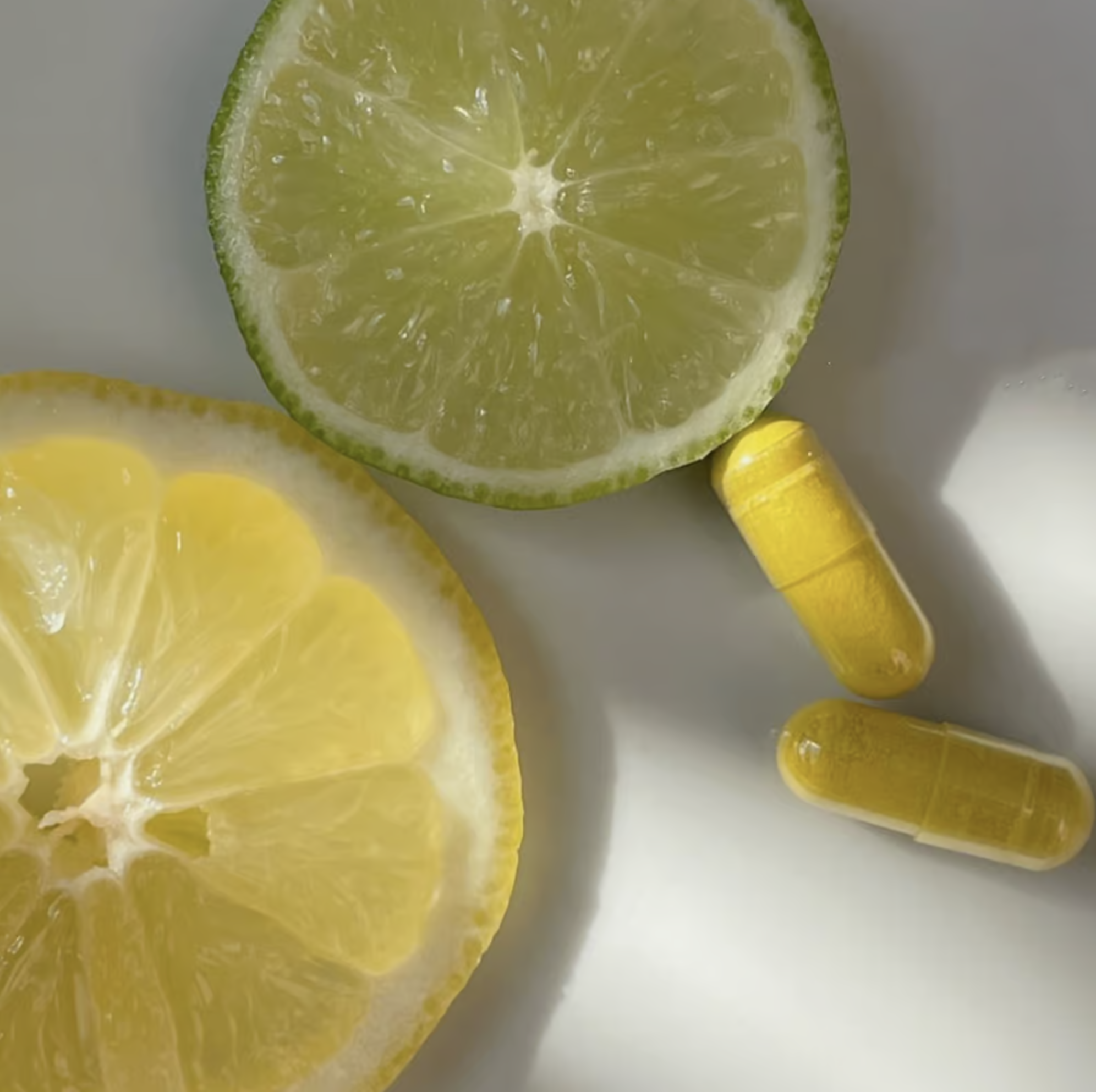 Two lemon slices, one green and one yellow, are placed next to three yellow capsules on a white surface.