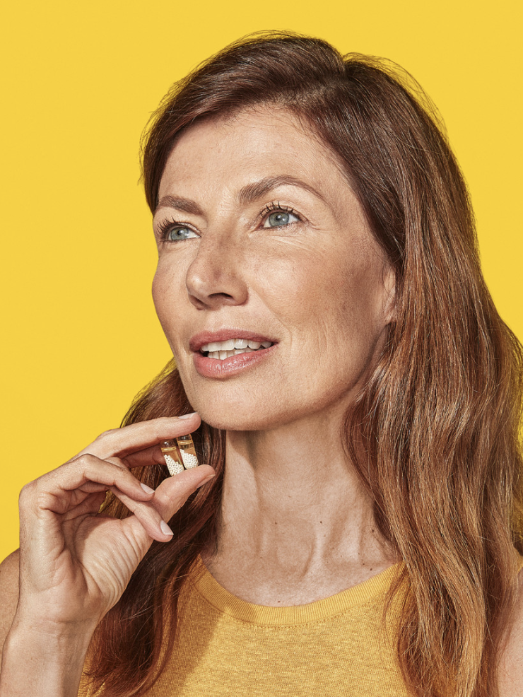 A woman with long brown hair and a yellow sleeveless top is holding her hand near her face against a yellow background.