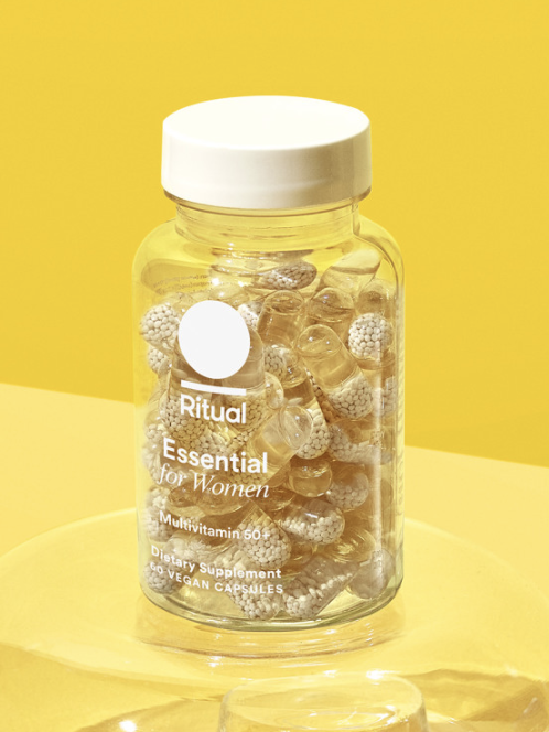 A clear bottle of Ritual Essential for Women vitamins with a white cap, placed on a glass surface against a yellow background. Text reads "Mint Essenced" with a leaf icon.