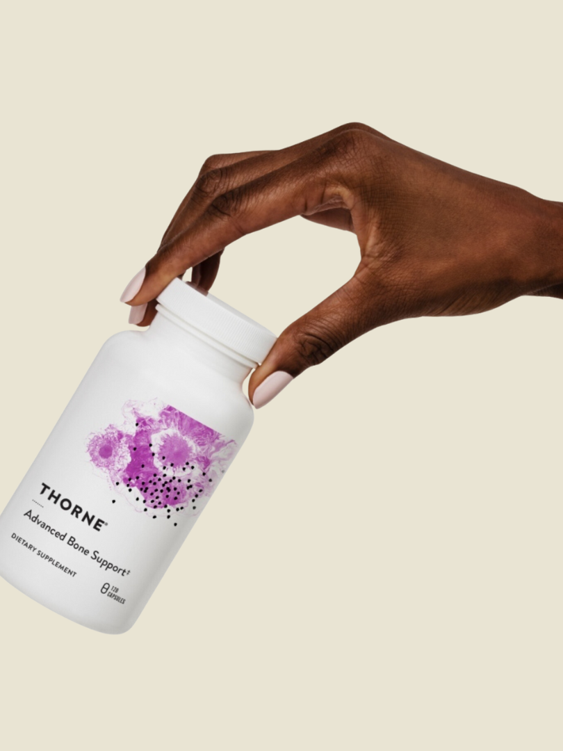 A hand holding a white bottle labeled "Thorne Advanced Bone Support." The bottle has a purple abstract design and contains dietary supplements.