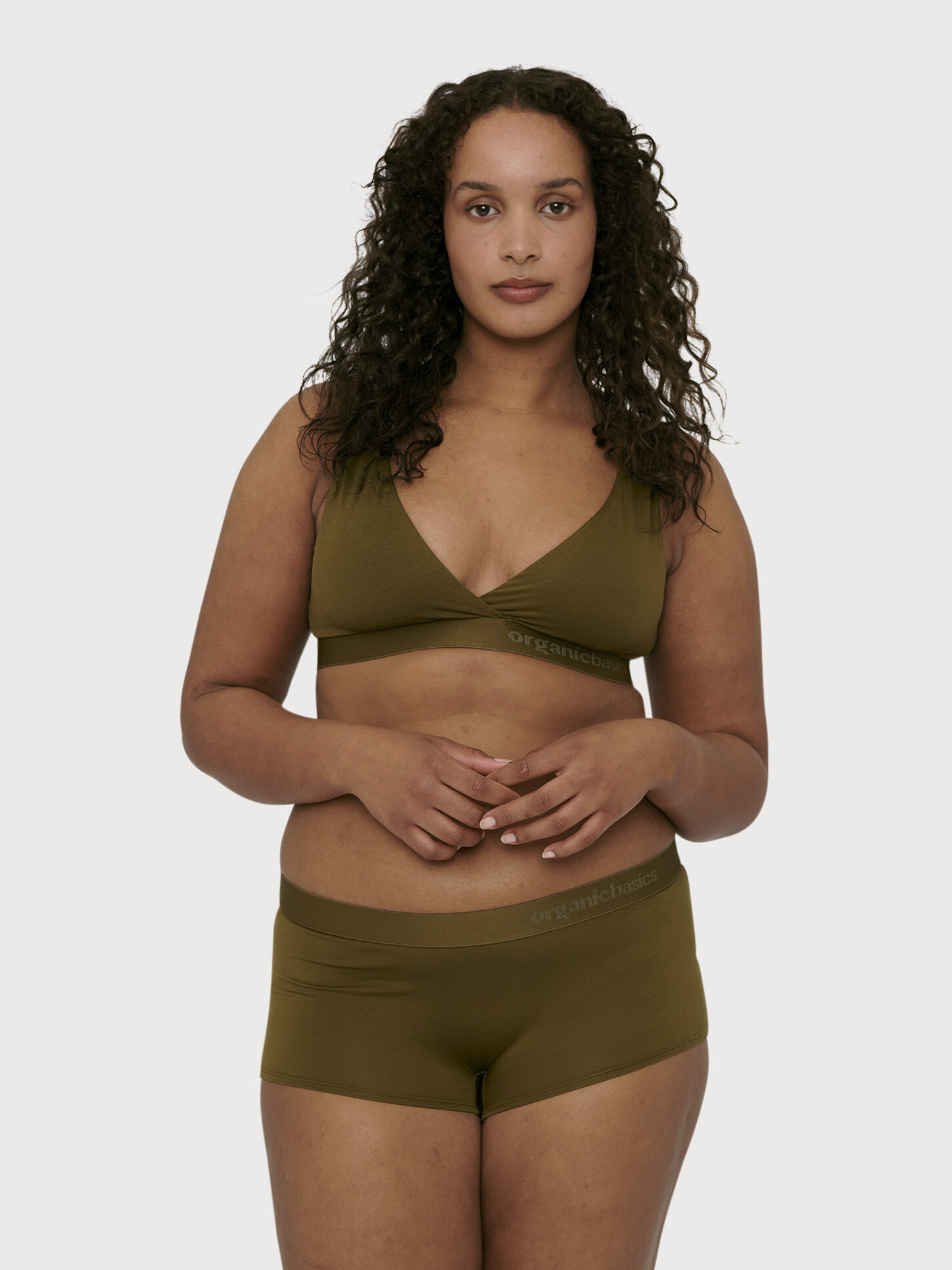 A woman with curly hair stands against a plain background, wearing an olive green bralette and matching boyshorts.