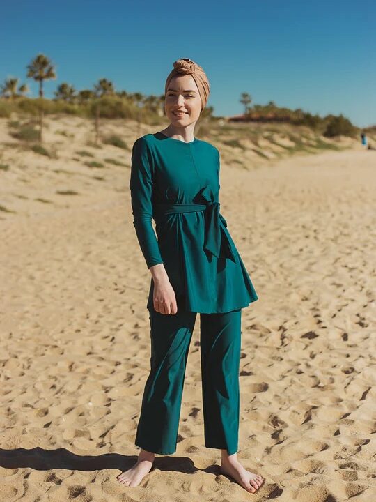 A person standing barefoot on a sandy beach wearing a teal outfit, with palm trees and a clear blue sky in the background.