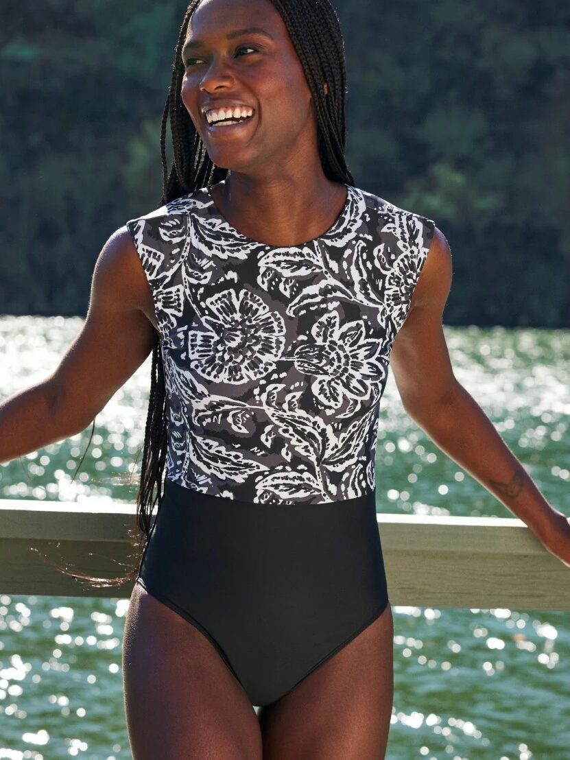 A person with long braided hair smiles while standing by water, wearing a black and white patterned swimsuit.