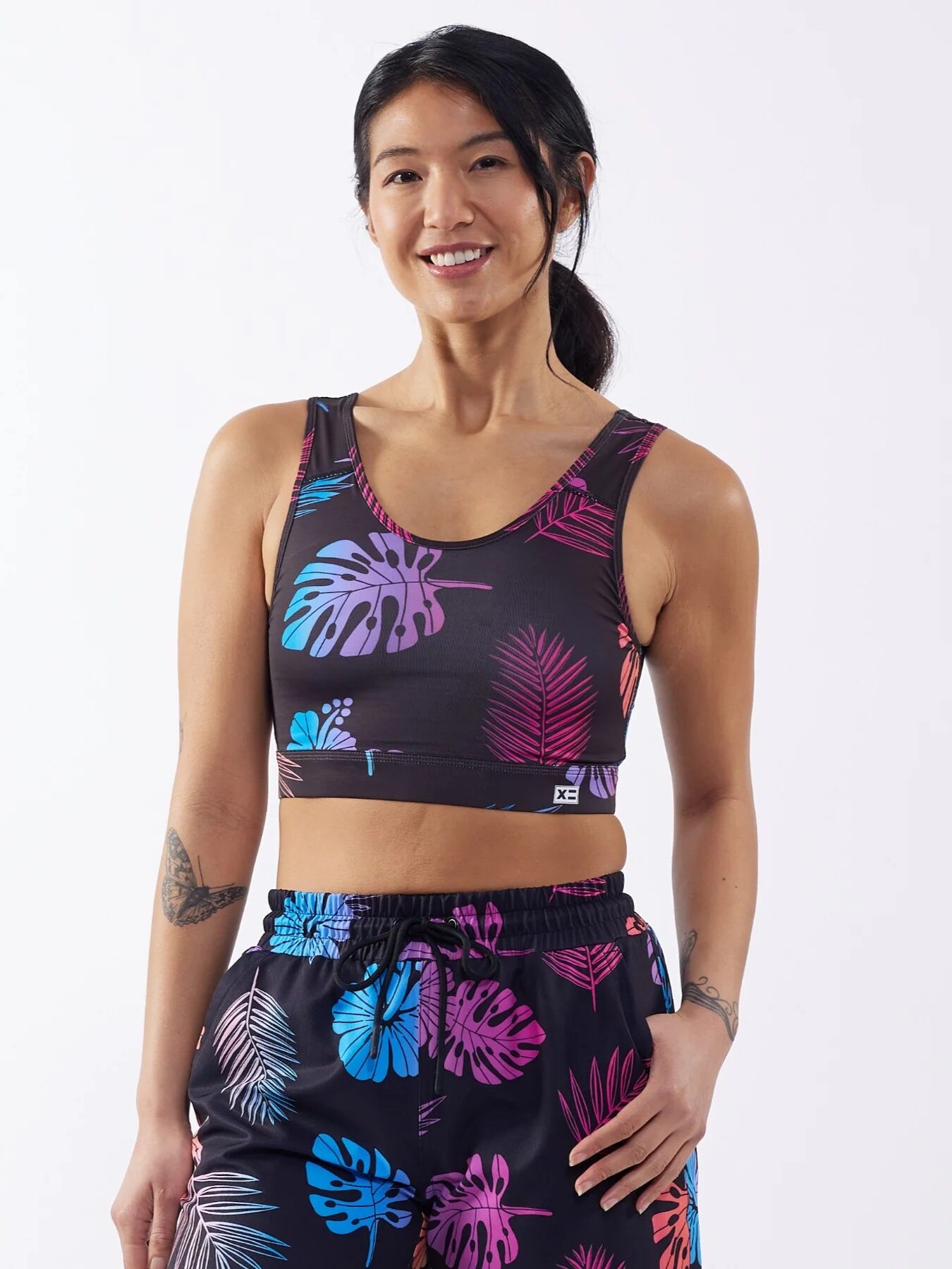 A woman is smiling and wearing a matching athletic outfit with a colorful tropical leaf pattern. She has a tattoo on her right forearm and her hair is tied back.