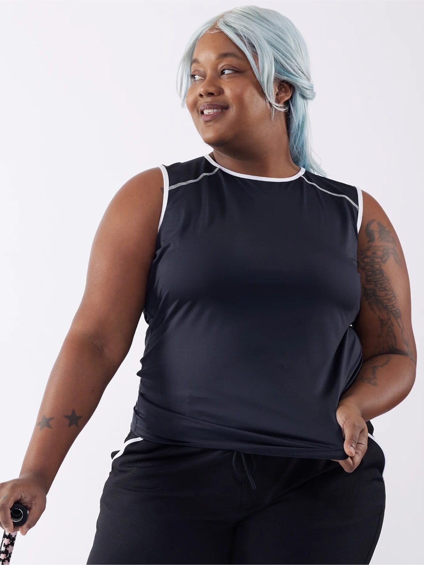 Person with light blue hair wearing a black sleeveless top and black pants, holding something in their left hand. They have tattoos on both arms and are looking to the side, smiling.