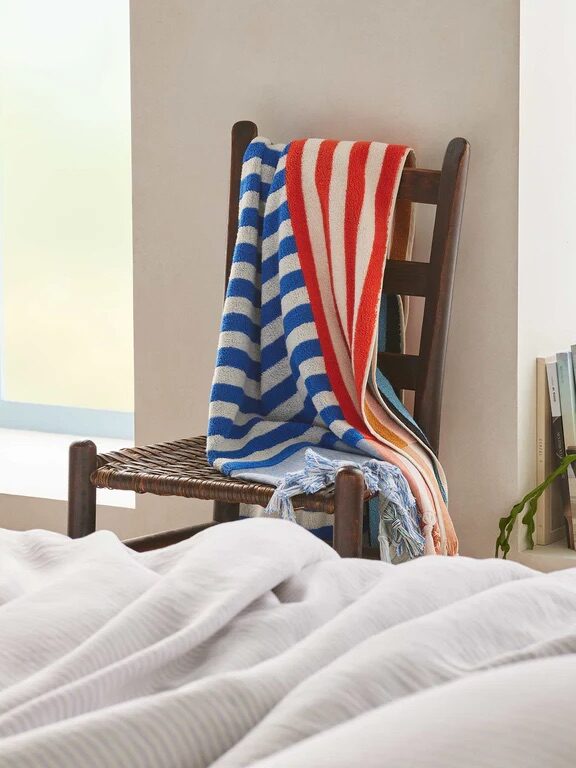 A wooden chair with a woven seat holds a striped blanket. A bed with white linens is in the foreground, and a plant and books are near the chair.