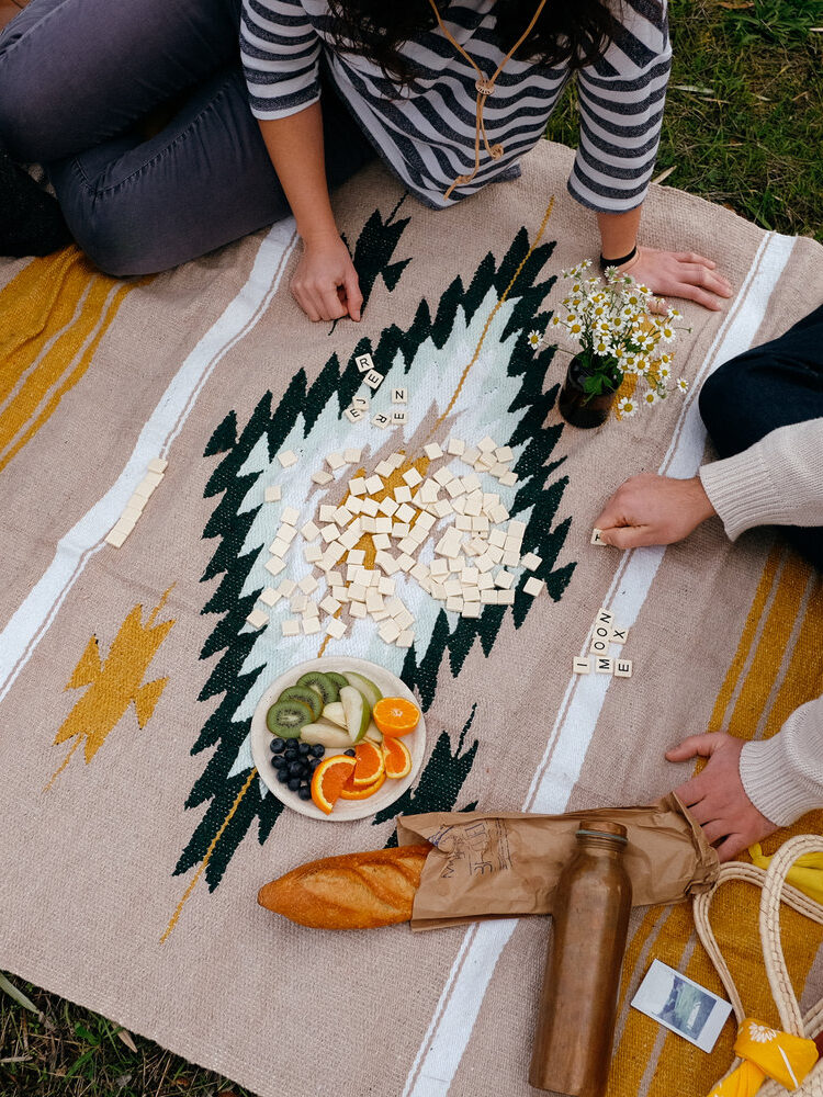 Three people sit on a patterned blanket outdoors, playing a board game with letter tiles. Snacks including fruit, bread, and a bottle of wine are arranged nearby.