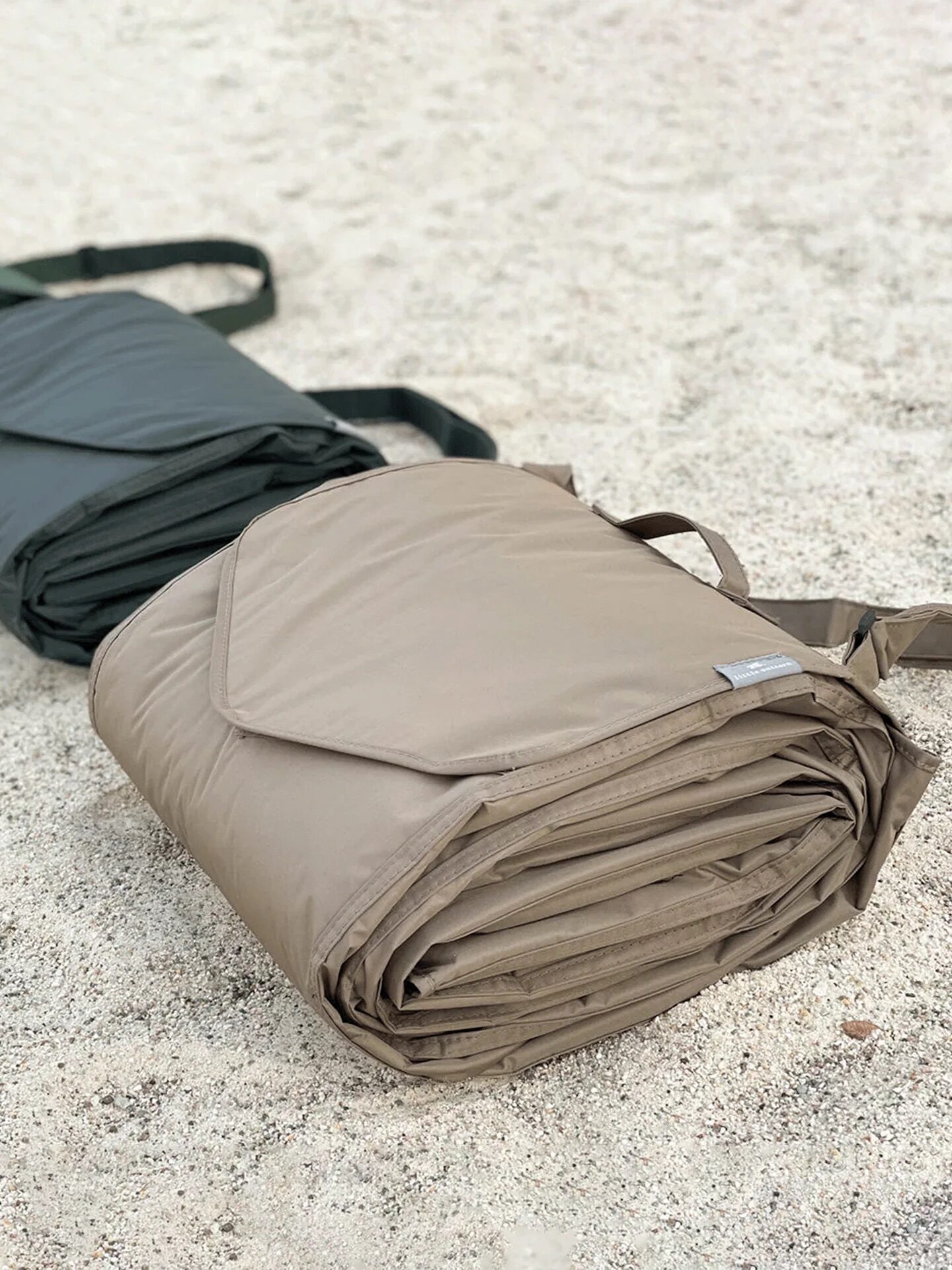 Three rolled-up sleeping bags, in green, dark green, and beige, are laid out on a sandy surface.