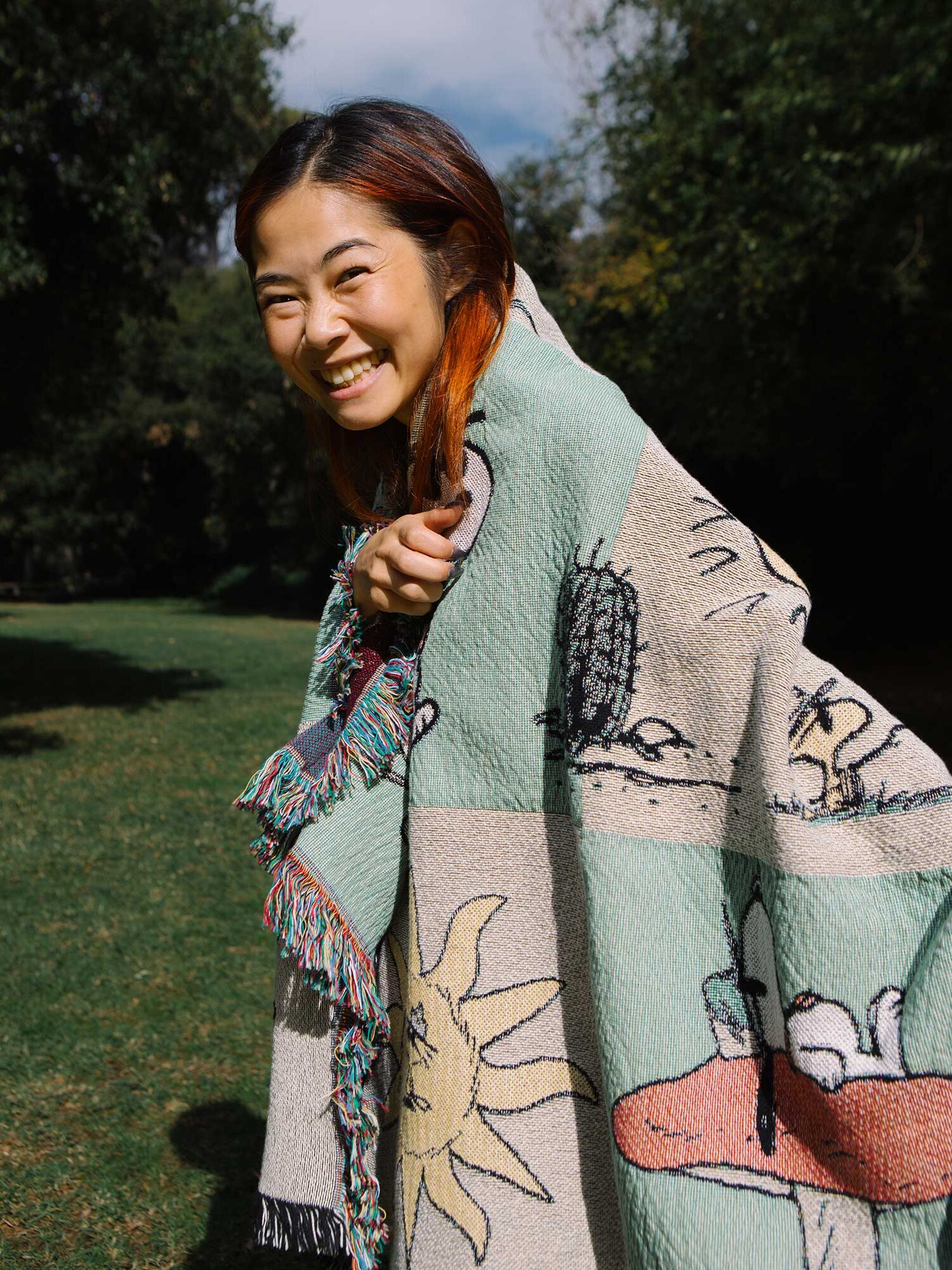 A person wrapped in a cartoon-patterned blanket smiles outdoors in a grassy area with trees in the background.