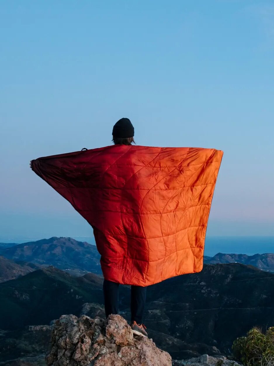 Person standing on a rocky peak overlooking mountains and sea during sunset, with an orange quilt wrapped around them.